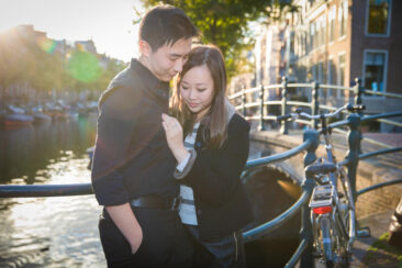 Couple photography, proposal and engagement photoshoot, portrait of an Asian man and woman posing by a sunny canal, both looking at the engagement ring of his fiancée, Amsterdam