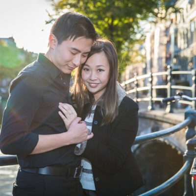 Couple photography, proposal and engagement photoshoot, portrait of an Asian man and woman posing by a sunny canal, the man is looking at the engagement ring of his fiancée, Amsterdam
