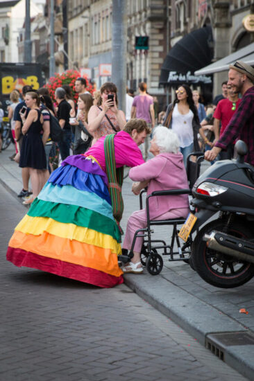 Journalistic photography, event photography, inclusive and inter-generational conversation between an old lady on a wheelchair and a young man dressed in a rainbow dress at Pink Saturday during Gay Pride, Amsterdam