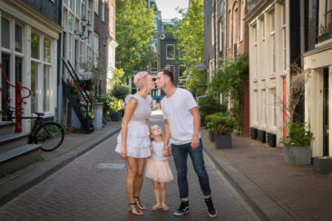 On-location family photoshoot in Amsterdam, children Photoshoot, together with the family for a fun photo session, happy family posing in a small street of old city centre Amsterdam, while the parents are kissing each other, the little girl looks up at them, imitating the kiss, Amsterdam