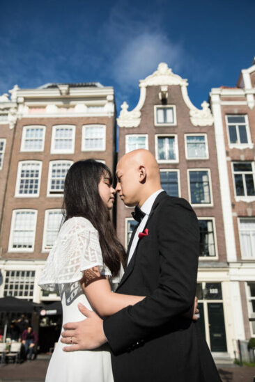 Couple photoshoot, loveshoot, engagement photoshoot, holiday photographer Amsterdam vacation photographer Amsterdam: smartly dressed Asian man and women hugging in front of typical old canal houses in Amsterdam old city centre