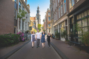 Family portrait photoshoot, parents and teenagers are walking together hand in hand in a small street of Amsterdam's old city centre with Westerkerk in the background