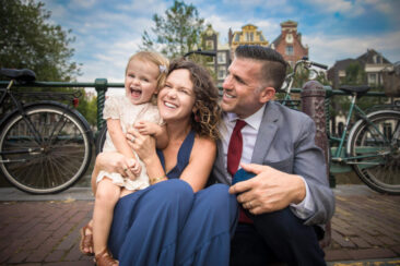 Familie fotosessie Amsterdam, On-location family photoshoot in Amsterdam, children Photoshoot, together with the family for a fun photo session, a happy little girl is standing laughing with her parents, sitting down on a bridge over Brouwersgracht, Amsterdam
