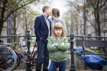 on-location family photoshoot Familie fotosessie Amsterdam, Family portrait photoshoot, a young girl is standing in front of her parents kissing in the background near a small canal in Amsterdam old city center