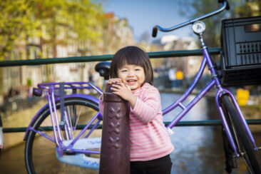 Familie fotosessie Amsterdam, On-location family photoshoot in Amsterdam, children Photoshoot, Family portrait, family photographer Amsterdam, portrait of a little girl laughing with a bike and a canal in thee background, Amsterdam old city centre